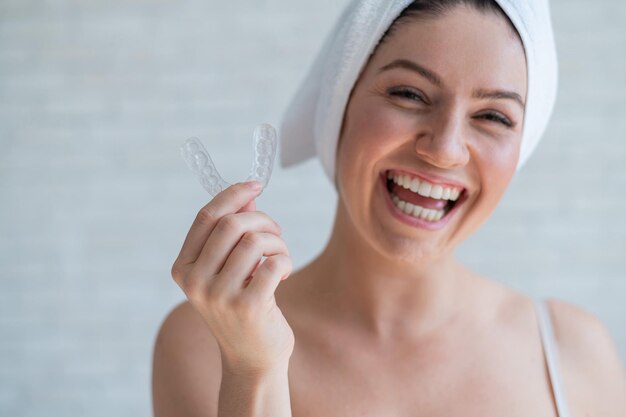 Une femme avec une serviette sur la tête tient un protège-dents blanchissant pour les dents La fille aligne ses dents à l'aide de dispositifs de retenue amovibles transparents de nuit Dispositif orthodontique pour un sourire parfait