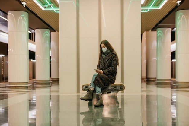 Une femme seule portant un masque médical pour éviter la propagation du coronavirus est assise à l'aide d'un smartphone sur le quai du métro. Une fille portant un masque chirurgical garde une distance sociale dans le métro.