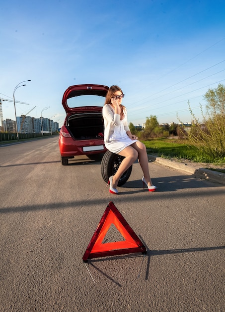 Femme sexy assise sur une roue de secours contre une voiture cassée et un panneau d'avertissement