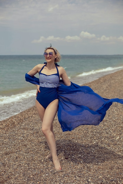 Femme sexy en maillot de bain bleu et lunettes de soleil marche sur la plage