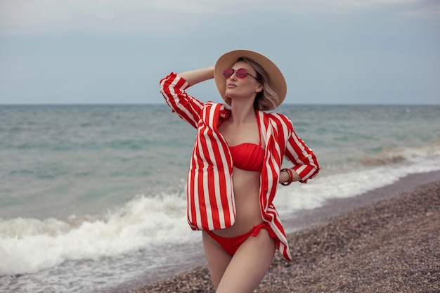 Une femme sexy en maillot de bain rouge, une veste rayée, des lunettes de soleil et un chapeau de paille se dresse sur la plage au bord de la mer en été