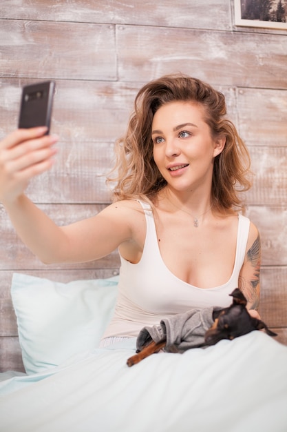 Femme sexy en pyjama prenant un selfie avec son chien au lit la nuit.