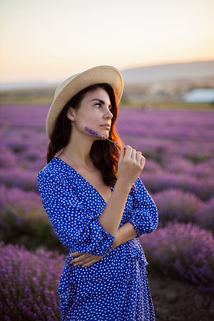 Une femme sexy vêtue d'une robe bleue et d'un chapeau de paille dans les champs de lavande en fleurs au coucher du soleil profite de la nature