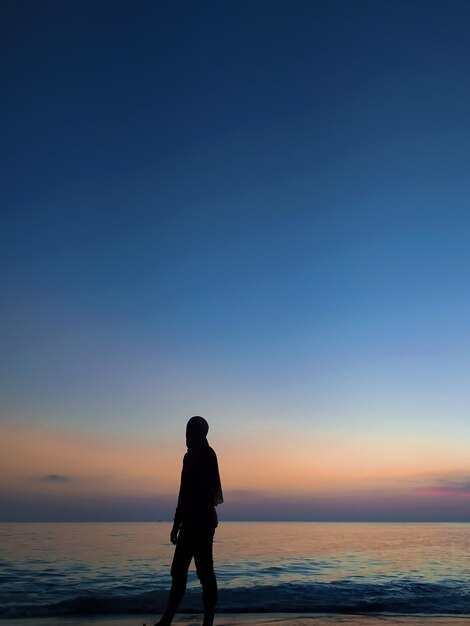 Une femme en silhouette debout sur la plage contre le ciel bleu au coucher du soleil