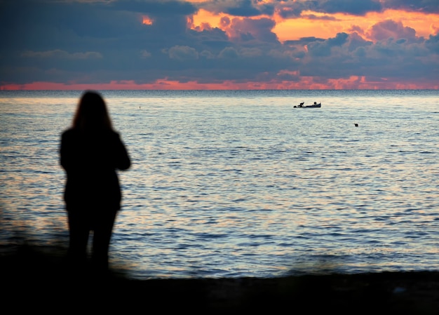 Photo la femme en silhouette observe la mer et le bateau dans les lumières du coucher du soleil. j'imagine que c'est la femme du pêcheur qui attend son retour.