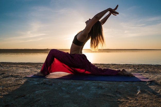 Femme silhouette sur la plage au coucher du soleil faisant du yoga asana. Entraînement d'échauffement naturel du matin