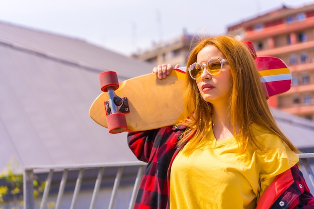 Femme de skateur dans une chemise jaune, une chemise à carreaux rouge et des lunettes de soleil, dans une pose avec la planche à roulettes tournée vers la gauche