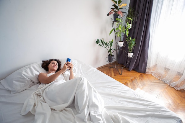 Femme avec smartphone au lit avec des draps blancs