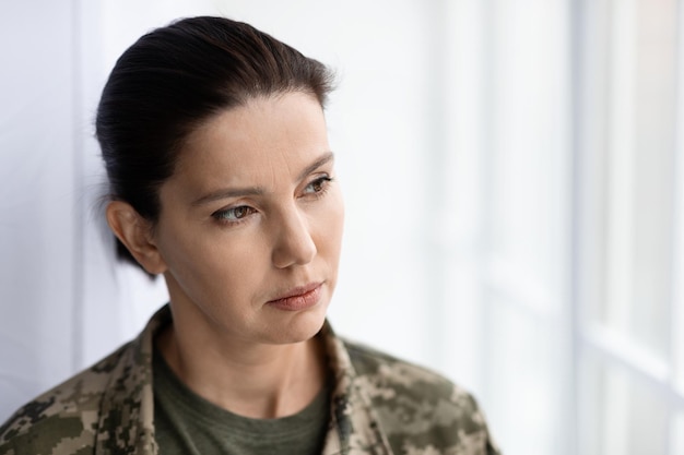 Une femme soldat réfléchie portant un uniforme de camouflage regardant par la fenêtre à la maison