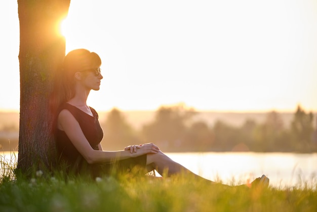 Femme solitaire assise seule sur une pelouse d'herbe verte penchée sur un tronc d'arbre sur la rive du lac lors d'une chaude soirée Solitude et détente dans le concept de la nature