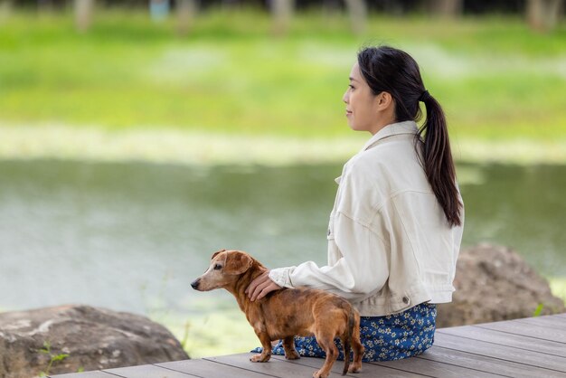 Une femme avec son chien dachshund est assise sur le sol en bois à côté du lac.