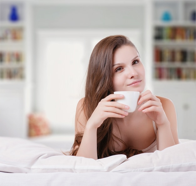 Femme sur son lit en souriant tout en tenant une tasse de café