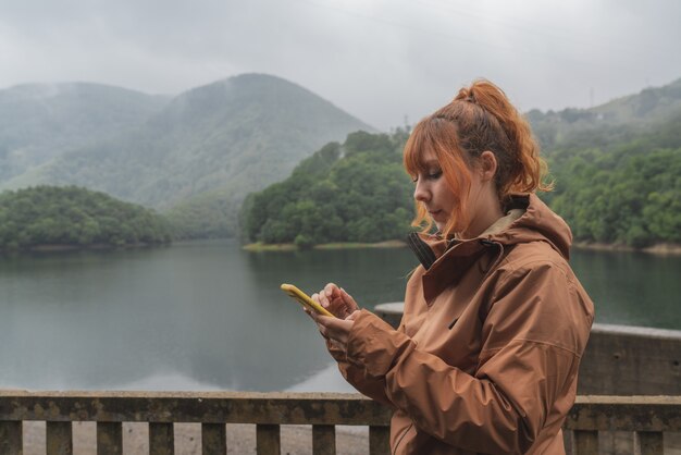 femme avec son smartphone devant un magnifique lac entouré de montagnes par temps nuageux