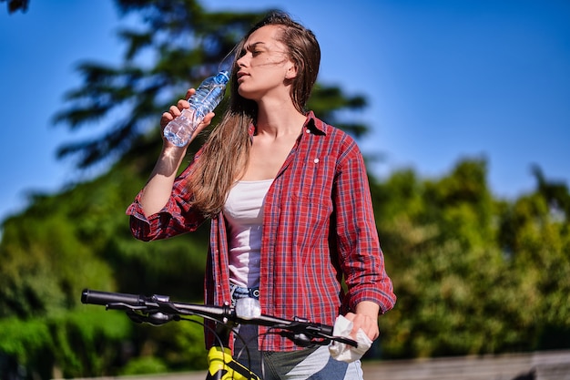 Femme souffrant de chaleur et de soif boit de l'eau fraîche et rafraîchissante pendant le vélo dans le parc en été