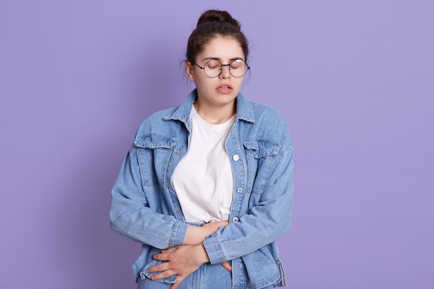 Femme souffrant de douleurs à l'estomac posant isolé sur un mur lilas, garde les yeux fermés, souffrant de douleurs au ventre, a l'air triste, vêtue d'une veste en jean et d'une chemise blanche.