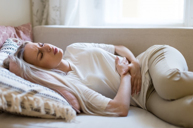 Femme souffrant de douleurs à l'estomac, ressentant des douleurs abdominales ou des crampes, allongée sur le canapé.