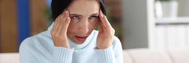 Une femme souffrant de maux de tête se tient la main dans les tempes tout en étant assise sur les symptômes de la migraine de la tête du canapé