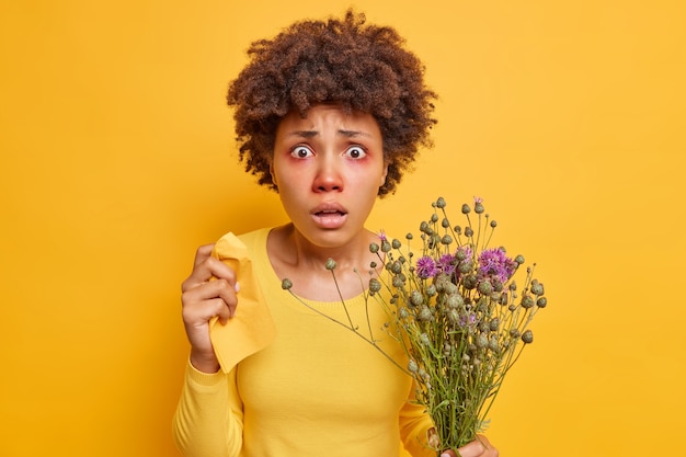 une femme souffre d'allergies saisonnières tient un bouquet de serviettes de fleurs sauvages a les yeux rouges qui démangent en réaction aux poses d'allergènes sur un jaune vif