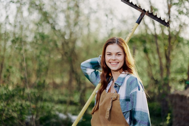 Une femme souriant magnifiquement et regardant la caméra un agriculteur en vêtements de travail et un tablier travaillant à l'extérieur dans la nature et tenant un râteau pour ramasser de l'herbe et du fourrage pour les animaux dans le jardin