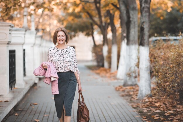 Une femme souriante de 50-55 ans se promène dans la rue en costume tenant un sac à l'extérieur en regardant la caméra
