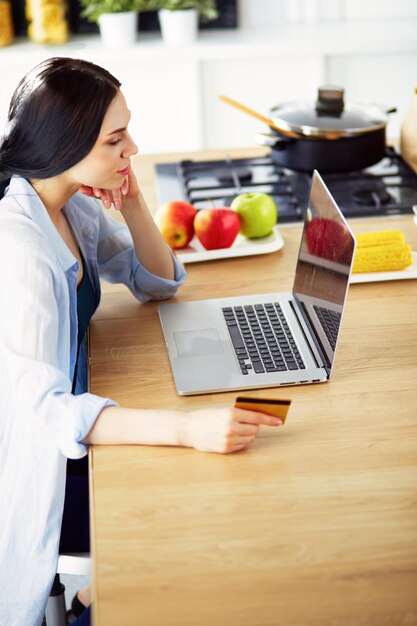 Femme souriante, achats en ligne à l'aide d'un ordinateur et d'une carte de crédit dans la cuisine