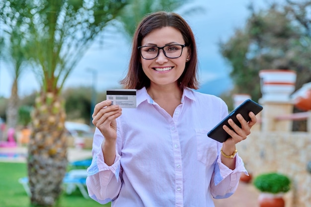 Femme souriante d'âge moyen avec carte de crédit et téléphone intelligent dans les mains en plein air