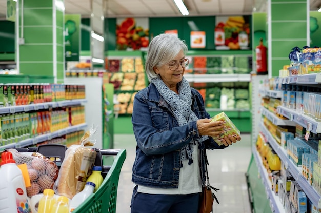 Femme souriante âgée avec des lunettes poussant un caddie dans le supermarché faisant des achats