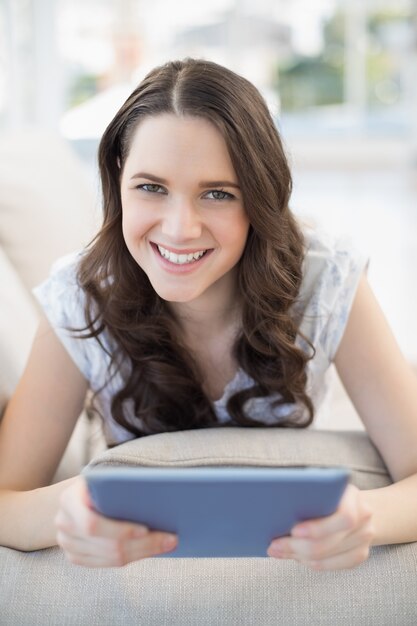 Femme souriante, allongée sur un canapé confortable tenant la tablette pc