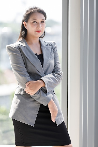 Une femme souriante asiatique en costume gris est debout dans la fenêtre du bureau. Concept confiant travaillant jeune femme d'affaires.