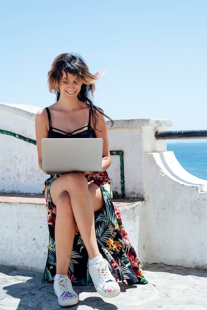 femme souriante assise sur un banc avec un ordinateur près de la mer