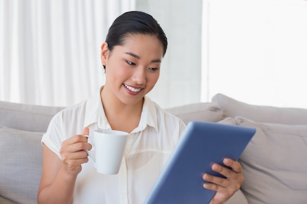 Femme souriante assise sur un canapé à l&#39;aide de tablet pc ayant un café