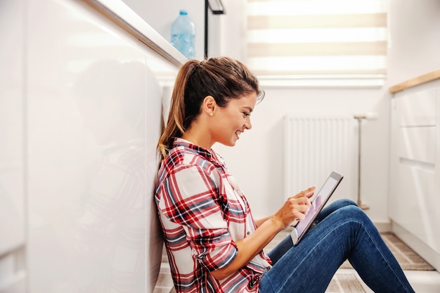 Femme souriante assise dans la cuisine sur le sol et à l'aide de tablette sur une pause.