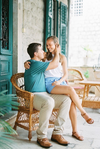 Photo femme souriante assise sur les genoux de l'homme dans une chaise en osier près de la vieille maison