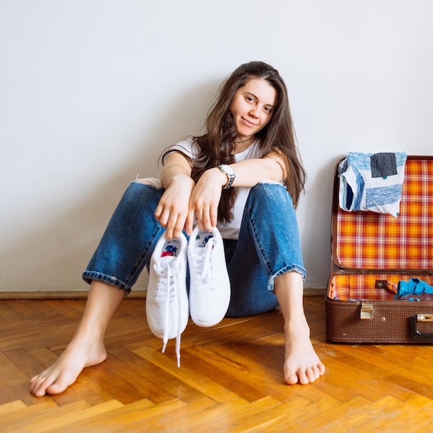 Femme souriante assise sur le sol près de la valise avec des vêtements emballés avant l'espace de copie du concept de voyage de voyage