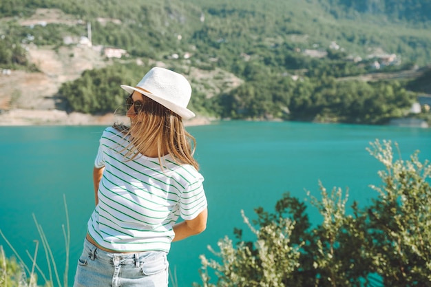 Femme souriante au chapeau et lunettes de soleil aux cheveux sauvages debout près du lac des montagnes sur fond