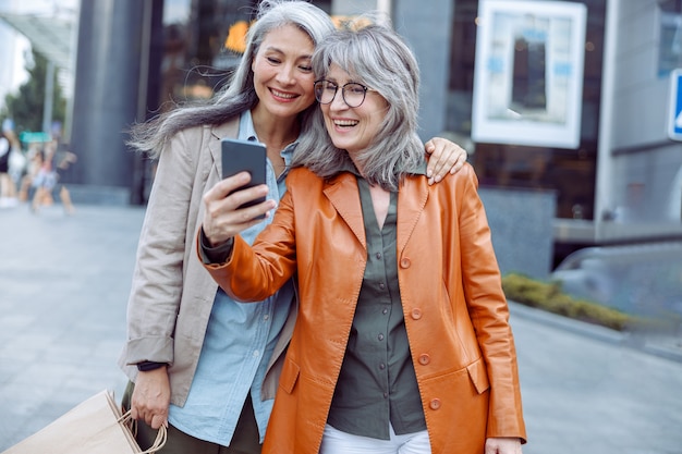 Une femme souriante aux cheveux argentés et son compagnon avec des sacs à provisions prennent un selfie dans une rue de la ville moderne