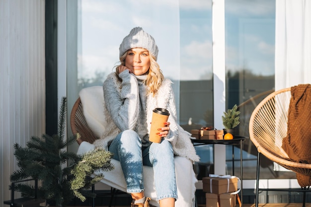 Femme souriante aux cheveux blonds en vêtements d'hiver avec une tasse de café assise près de la maison de campagne en hiver