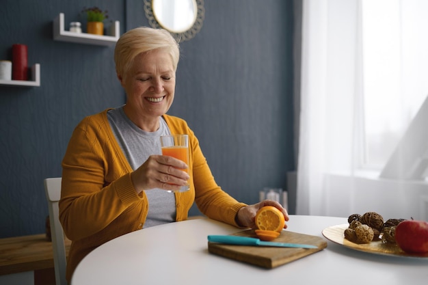 Femme souriante aux cheveux gris tenant une tasse en verre avec du jus d'orange. Oranges fraîchement cueillies sur table