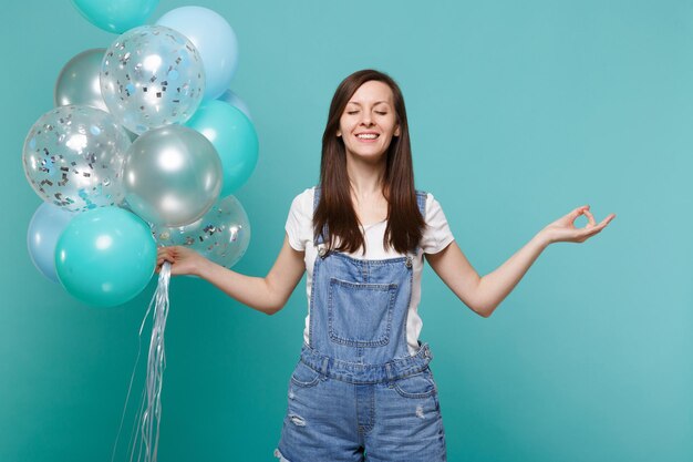 Femme souriante aux yeux fermés, se tenir la main dans un geste de yoga, se détendre en méditant, tenant des ballons à air colorés isolés sur fond bleu turquoise. Fête d'anniversaire, concept d'émotions de personnes.