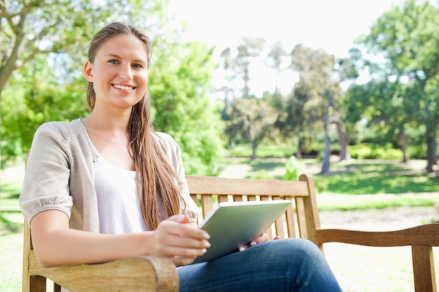 Femme souriante sur un banc dans le parc avec son ordinateur tablette