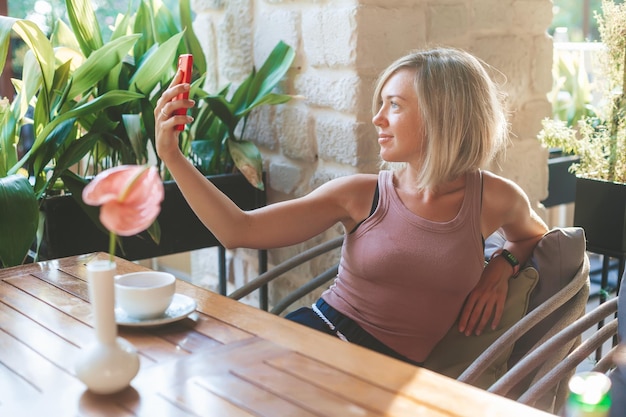 Femme souriante blonde faisant selfie sur un téléphone rouge tout en étant assis dans un café confortable