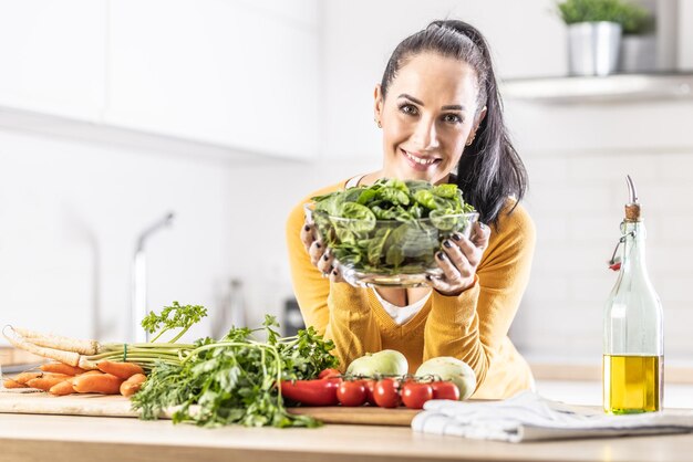 Femme souriante avec un bol plein de feuilles d'épinards avec des légumes frais et de l'huile d'olive dans sa cuisine