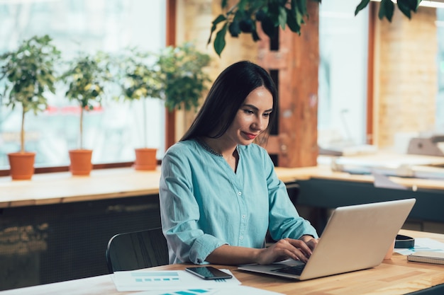 Femme souriante calme utilisant un ordinateur portable pour travailler tout en étant assise à la table