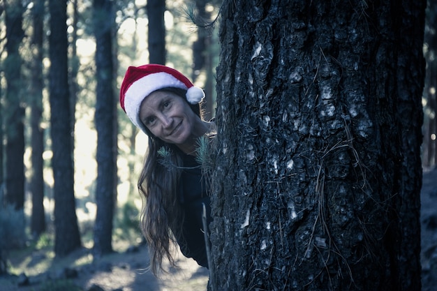 Femme souriante avec chapeau de Noël se penchant derrière un tronc de pin en forêt avec la lumière du soleil derrière