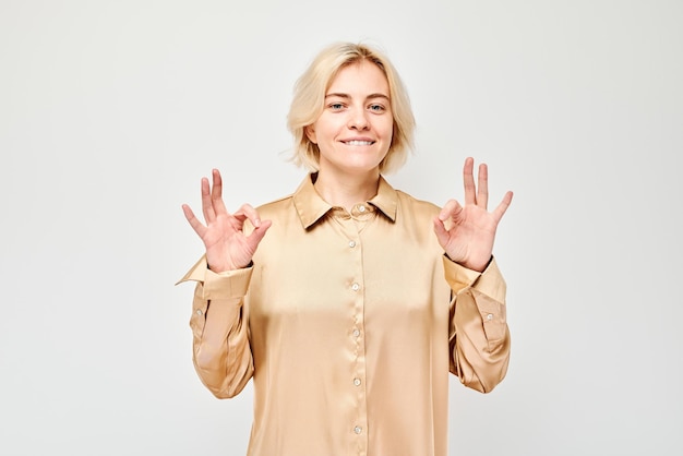 Une femme souriante en chemise beige fait un signe OK avec les deux mains sur un fond blanc