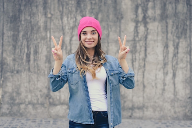 Femme souriante en chemise de jeans et chapeau rose montrant le signe de la victoire mur gris