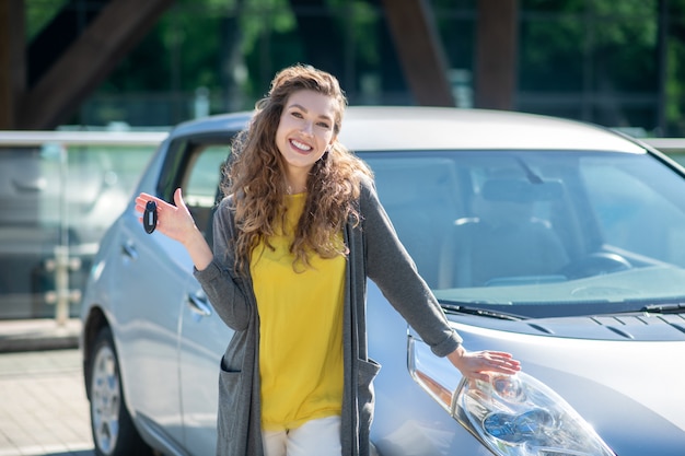 Femme souriante avec des clés debout près de la voiture grise