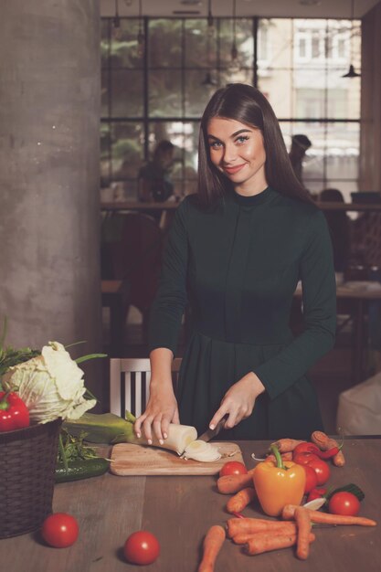 Une femme souriante coupe un oignon avec un couteau à la table avec beaucoup de légumes