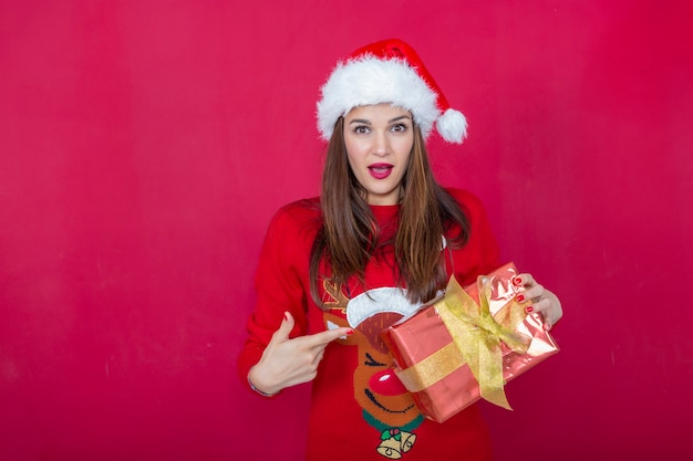 femme souriante dans un bonnet de Noel et pull renne de Noël pointant vers un cadeau dans sa main