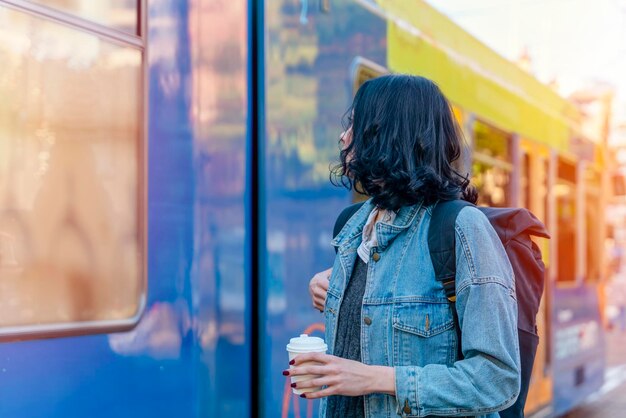 femme souriante dans une veste en jean buvant du café et attendant un tram à l'arrêt photo de style de vie